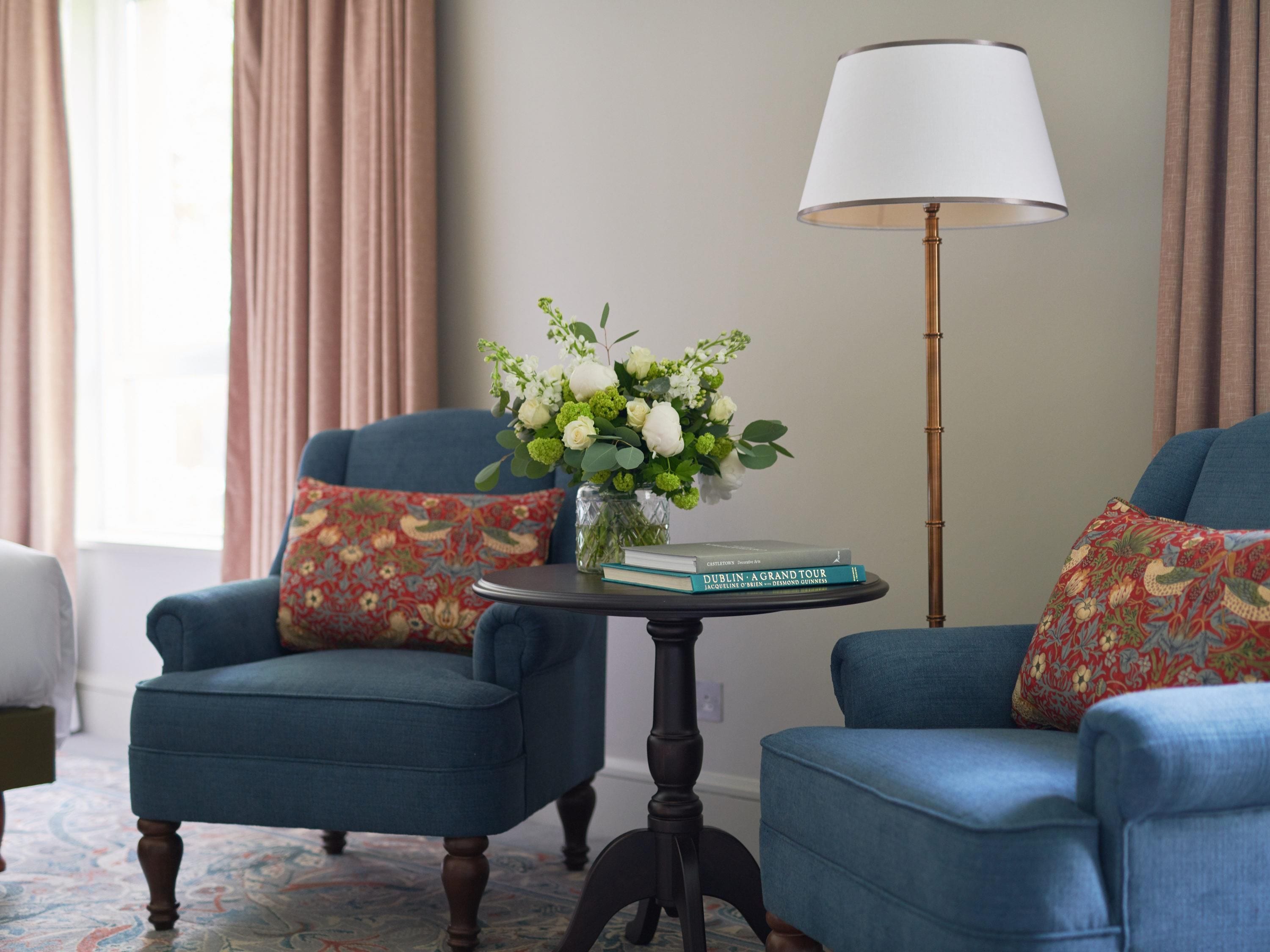 A room with two blue armchairs, a small round table with a book and a vase of flowers, and a floor lamp.