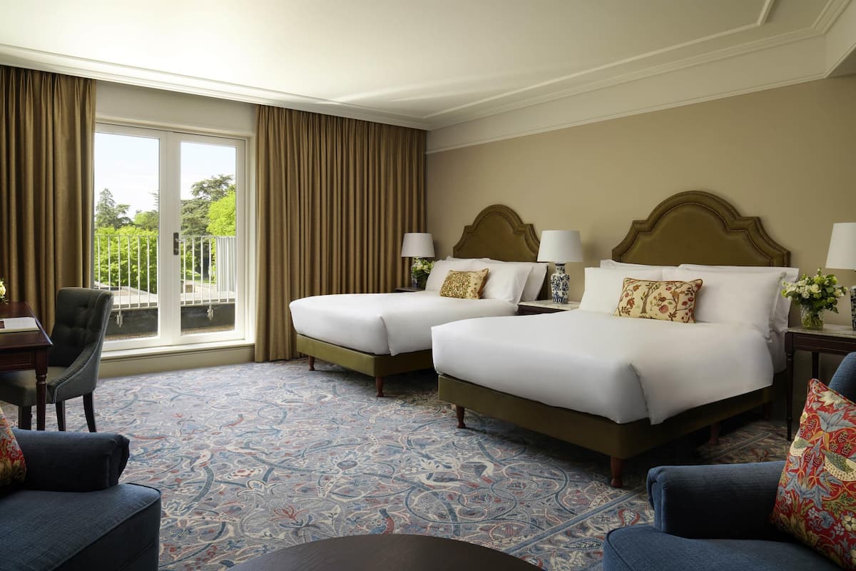Une chambre d’hôtel avec deux lits, un bureau et une chaise. Il y a une grande fenêtre avec des rideaux et une vue sur la verdure à l’extérieur.