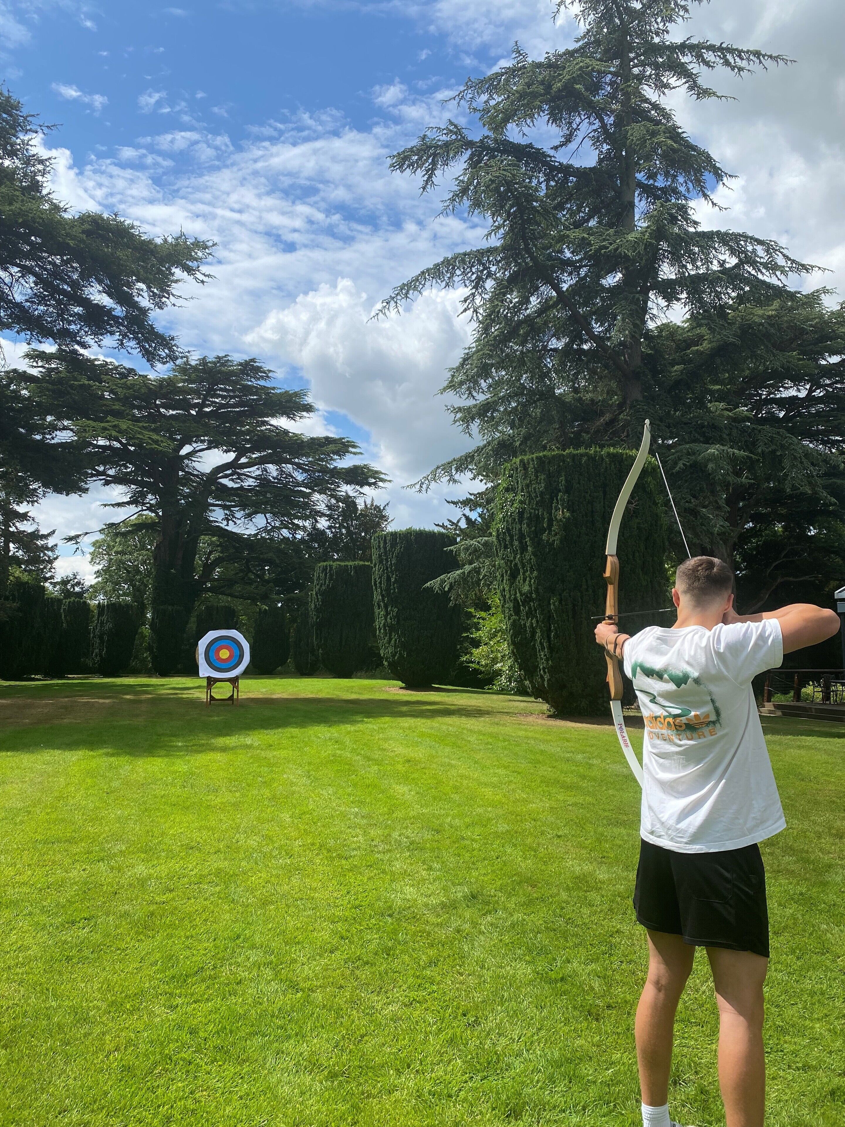 A person aiming an arrow at a target in a grassy area with trees in the background.