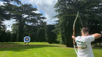 A person aiming an arrow at a target in a grassy area with trees in the background.