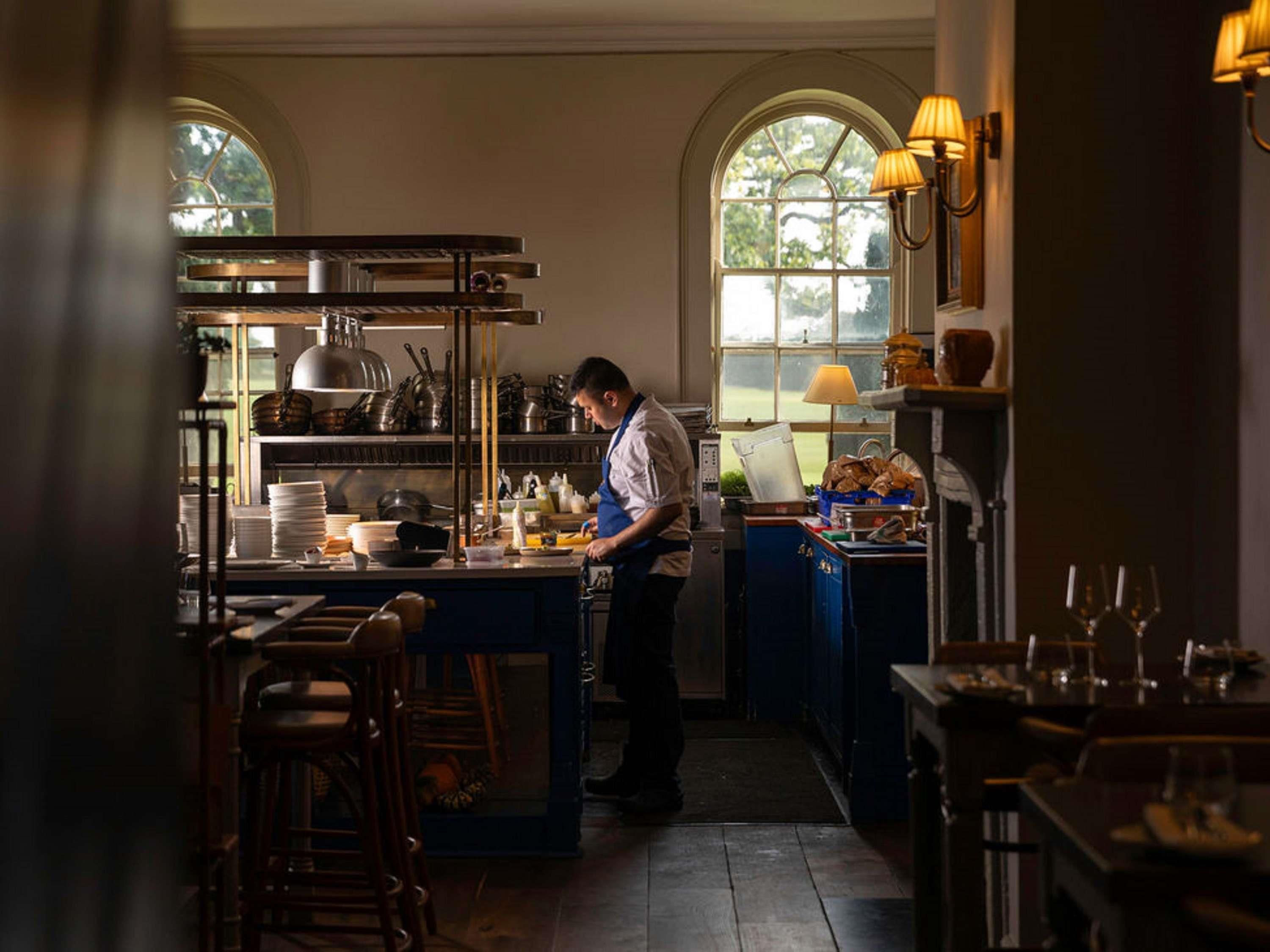 A chef preparing food in a well-lit kitchen with large windows, a bar area, and a dining space with tables and chairs.