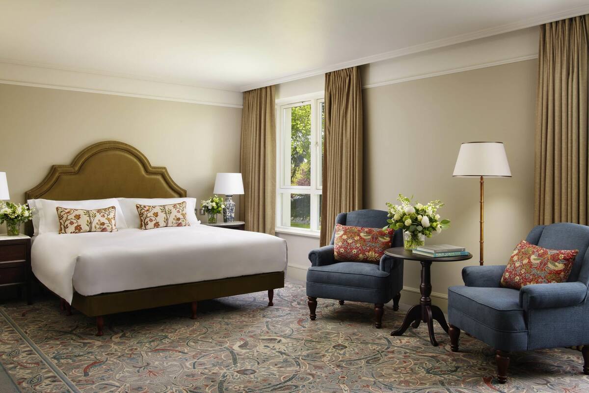 Une chambre d’hôtel avec un grand lit, deux fauteuils, une petite table et une lampe sur pied.