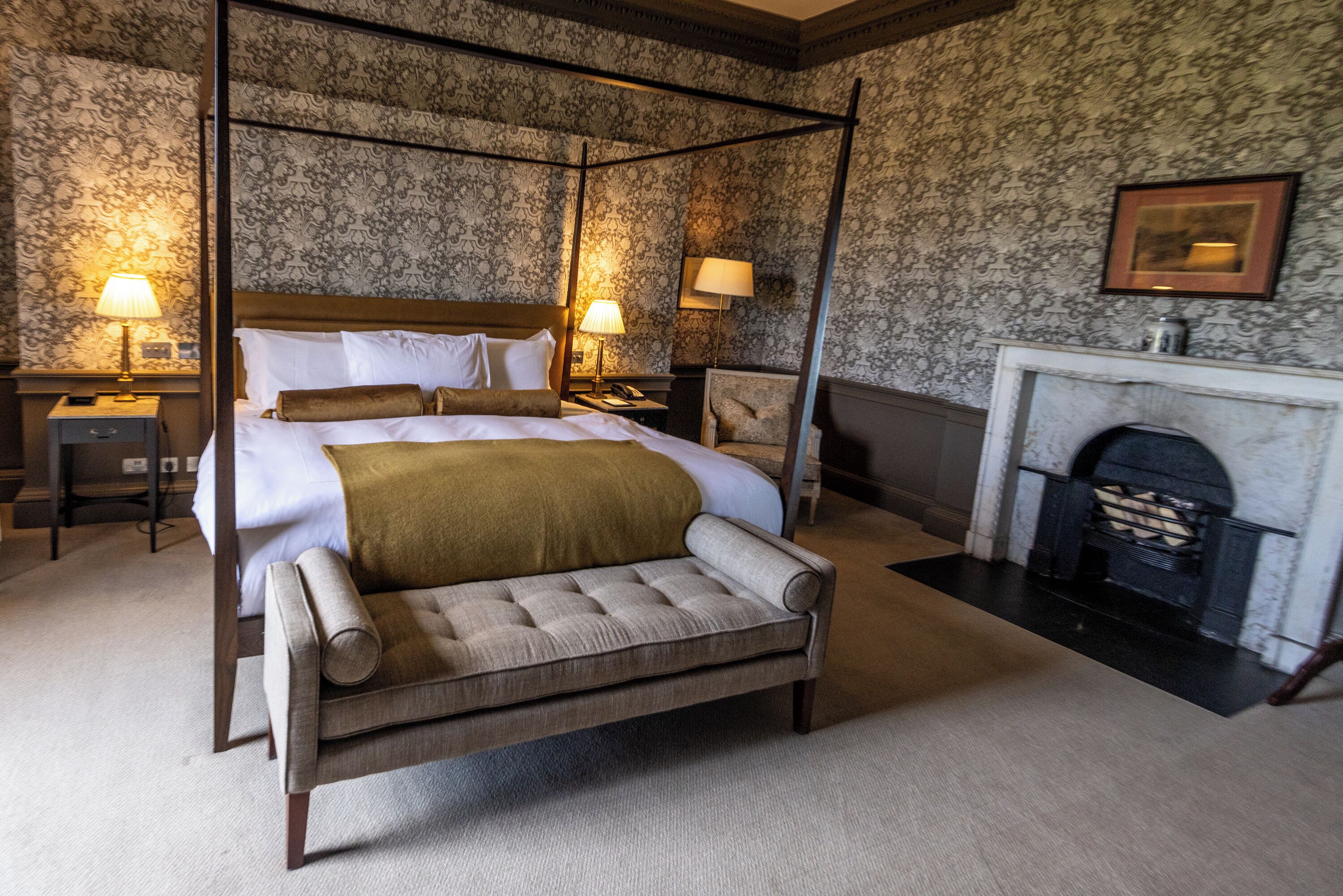 A four-poster bed with a headboard, two bedside tables with lamps, a chair, and a fireplace.