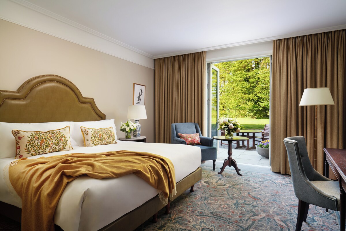 A hotel room with a large bed, a desk, a chair, and a view of a garden through an open door.