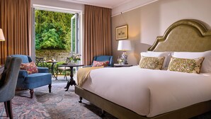 A hotel room with a large bed, two chairs, a table, and a view of greenery outside.