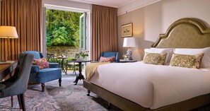 A hotel room with a large bed, two chairs, a table, and a view of greenery outside.