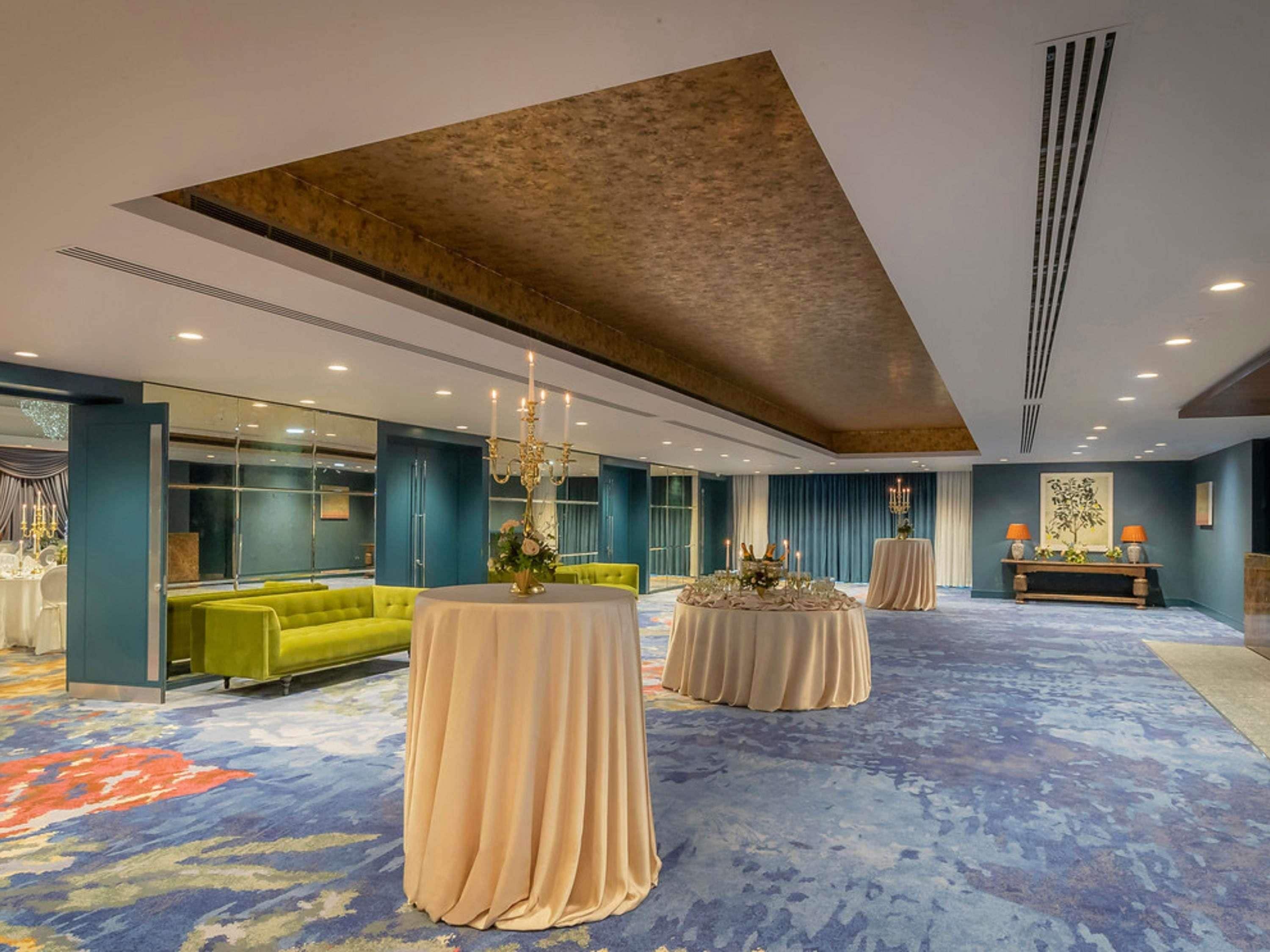 A spacious hotel lobby with a high ceiling, a green sofa, round tables, and a blue carpet.
