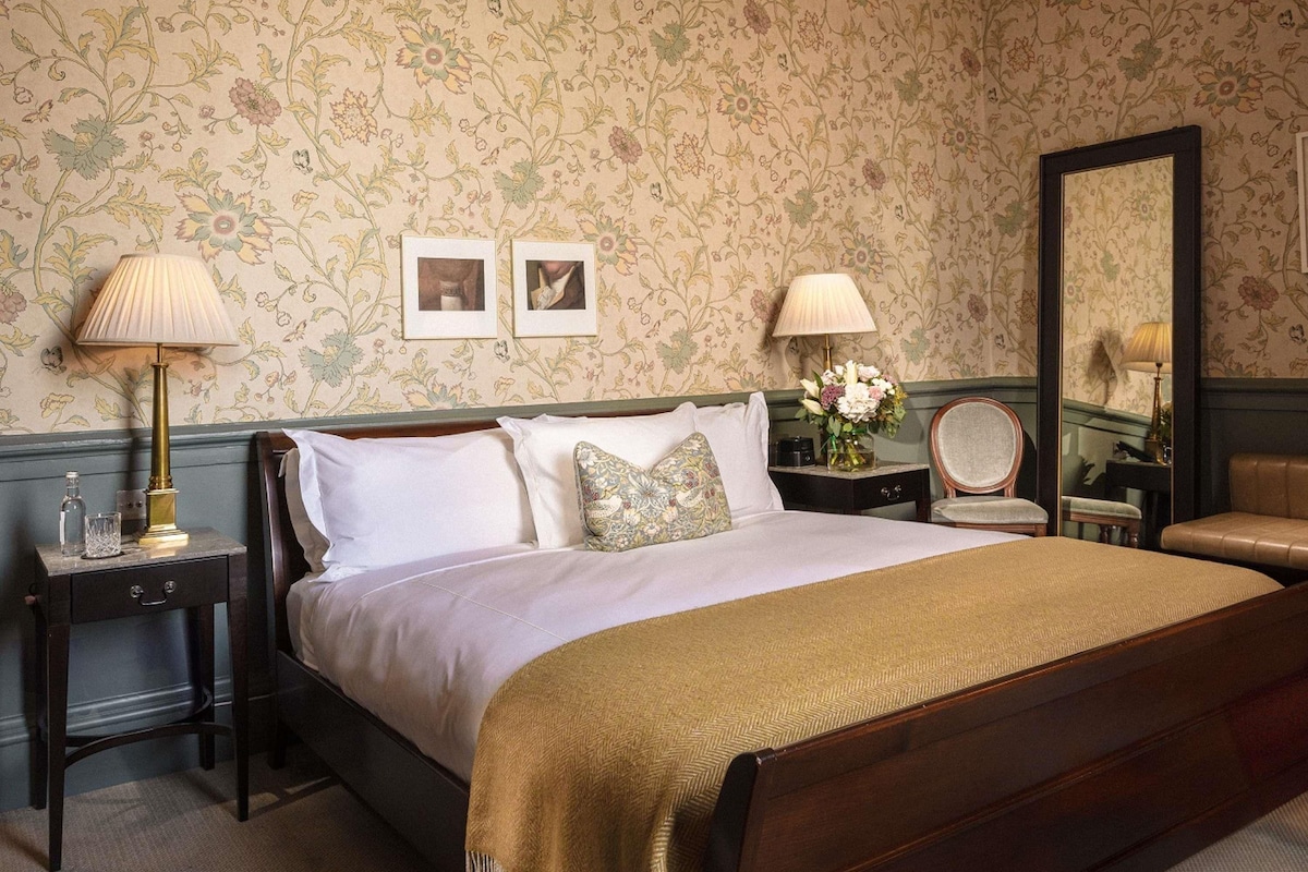 A neatly arranged bedroom with a large bed, two bedside tables, a mirror, and floral wallpaper.