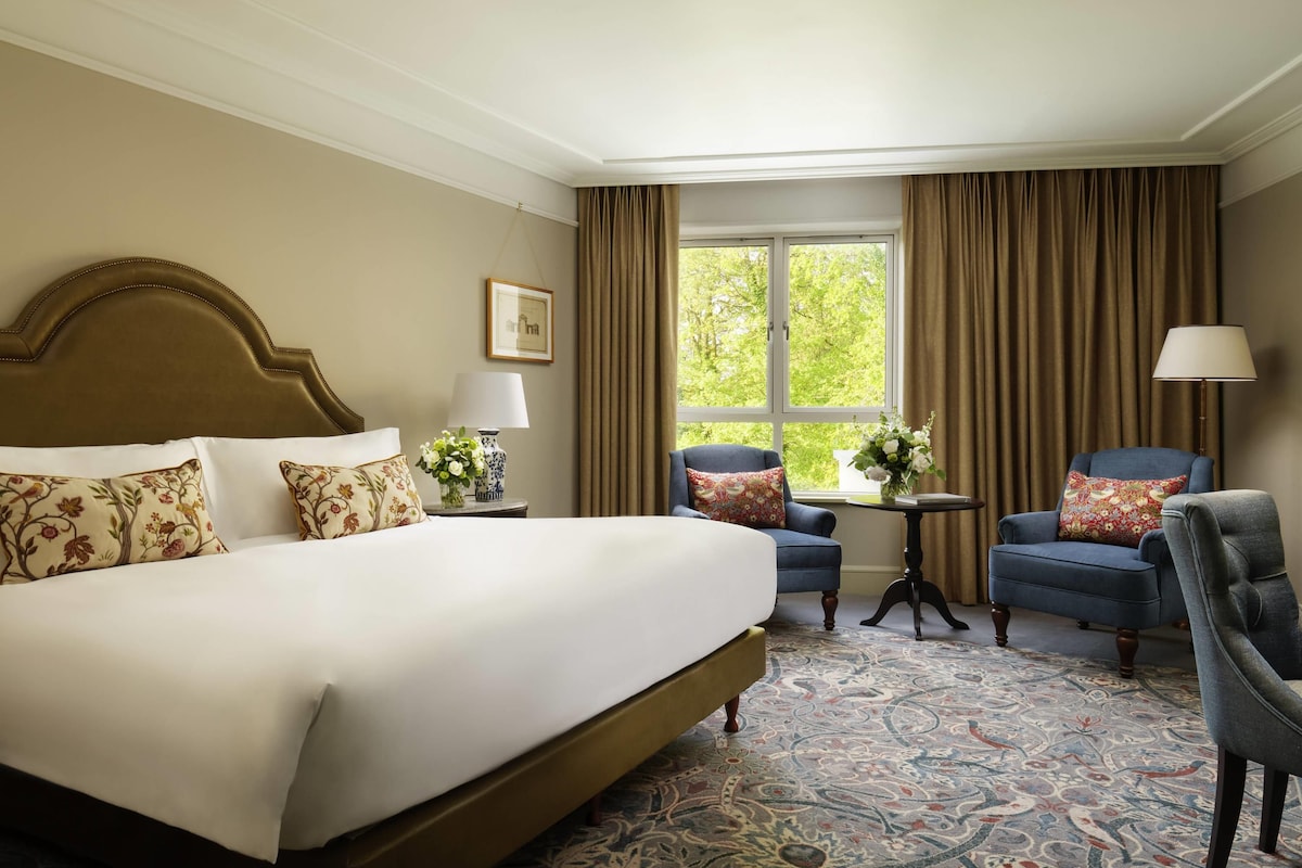 A hotel room with a large bed, two armchairs, a small table, and a window with curtains.