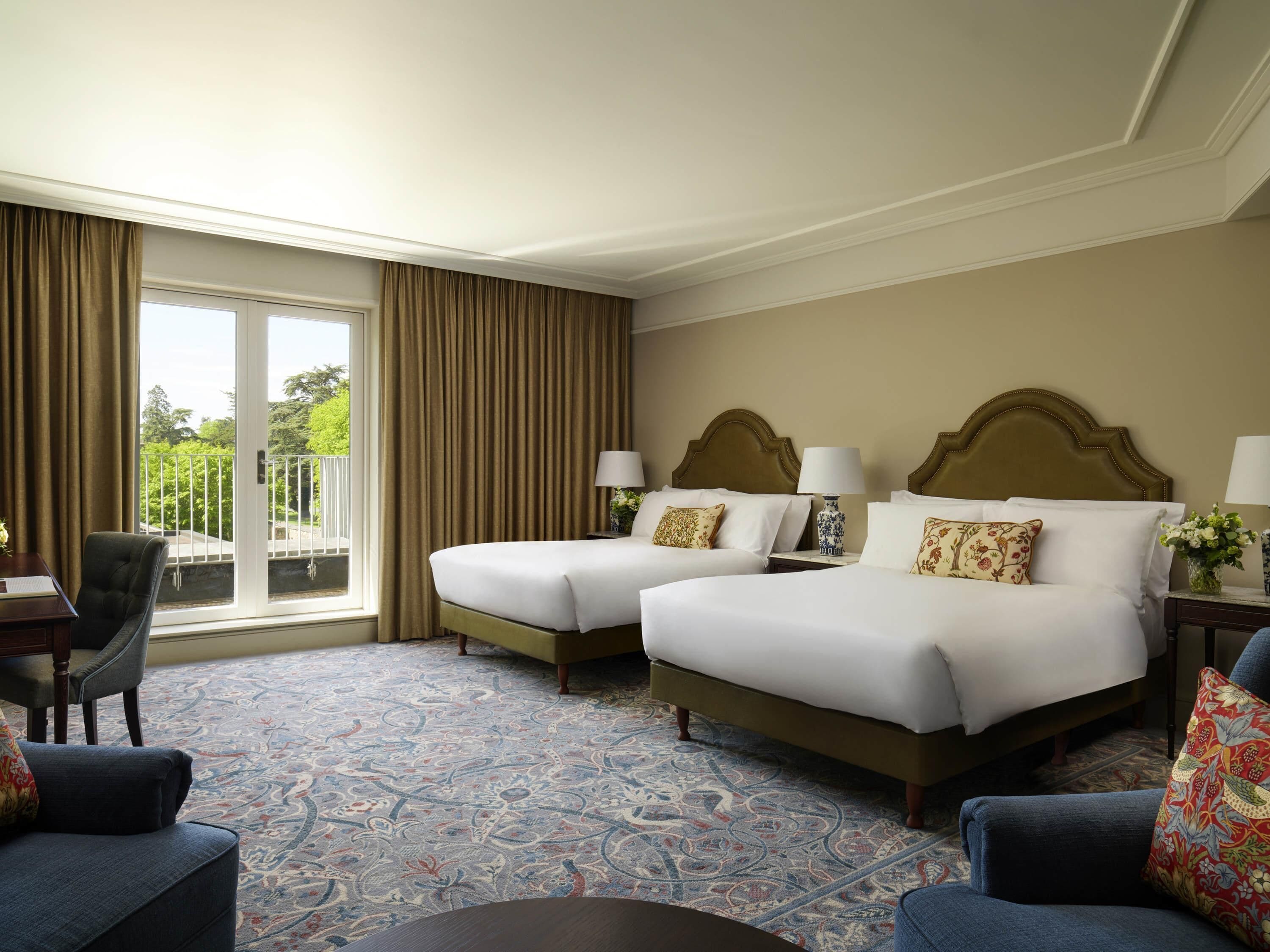 A hotel room with two beds, a desk, a chair, and a balcony with curtains.