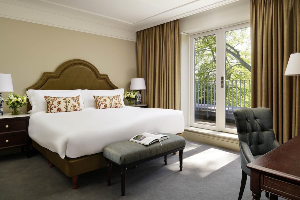 A hotel room with a large bed, a desk, a chair, a window with curtains, and a view of greenery outside.