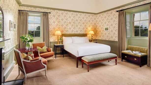 A hotel room with a bed, two bedside tables, a chair, a bench, and a window with curtains.
