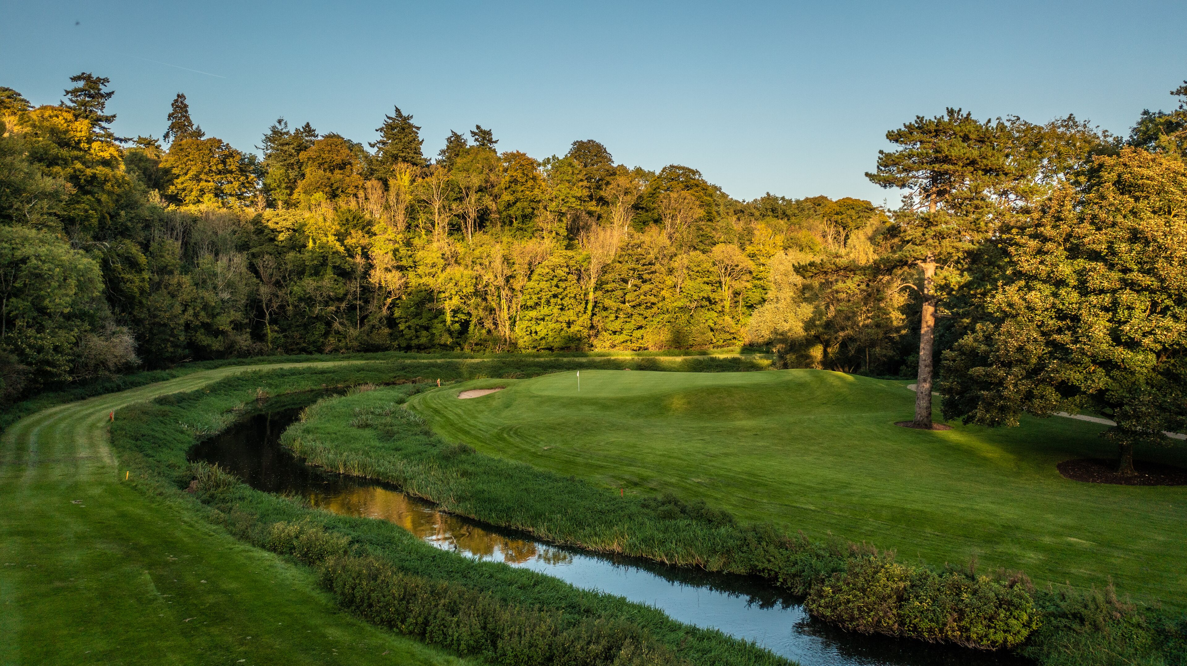 A golf course with a water feature, surrounded by dense trees and a clear sky.