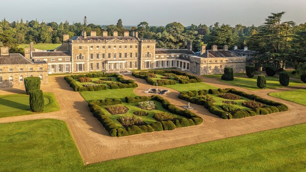 A large, historic building with a formal garden layout, including geometrically shaped hedges and a central fountain.