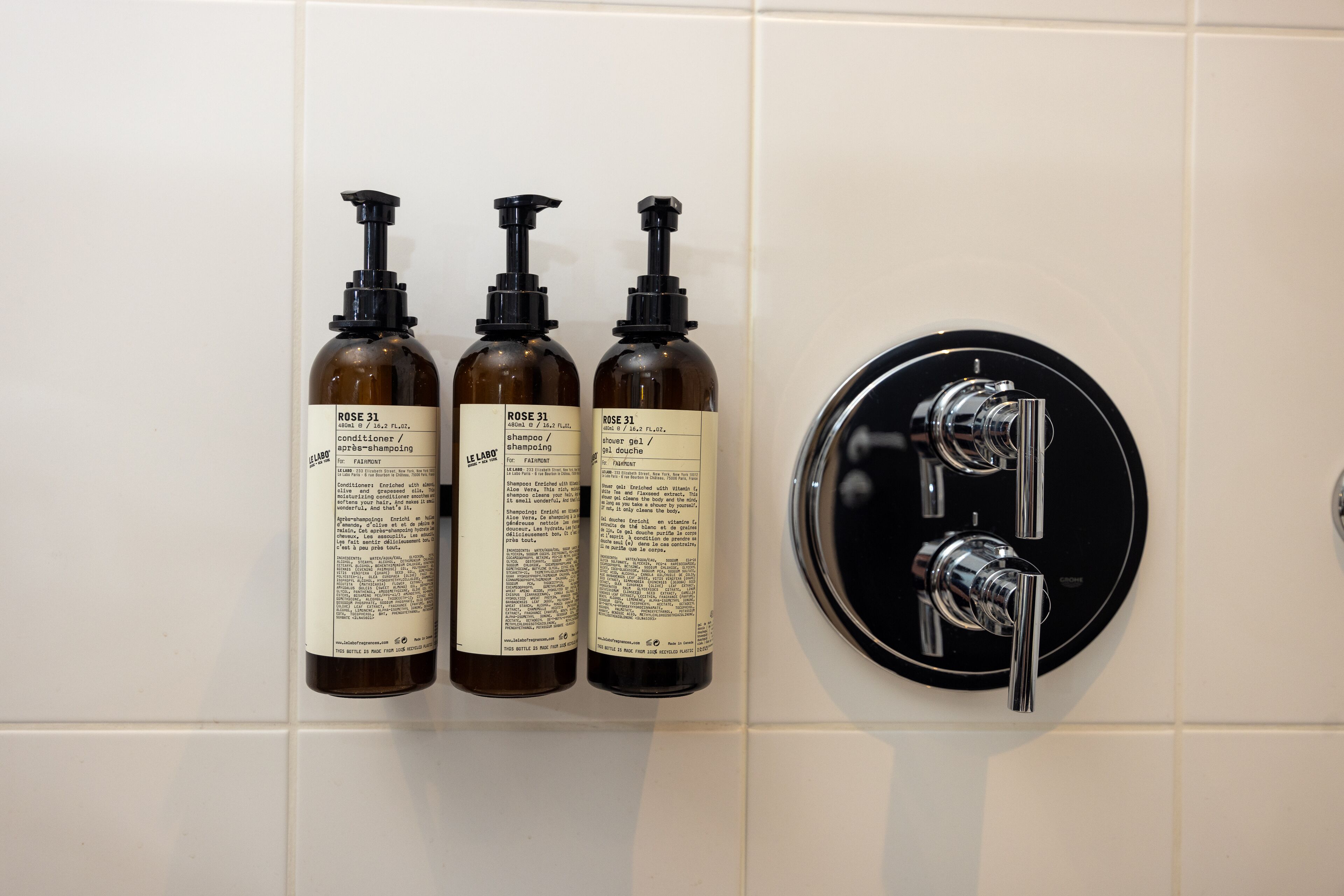 Three bottles of 'Rose 31' shampoo and conditioner, and a black faucet with two handles on a tiled wall.