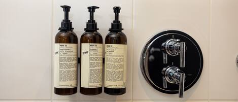 Three bottles of 'Rose 31' shampoo and conditioner, and a black faucet with two handles on a tiled wall.
