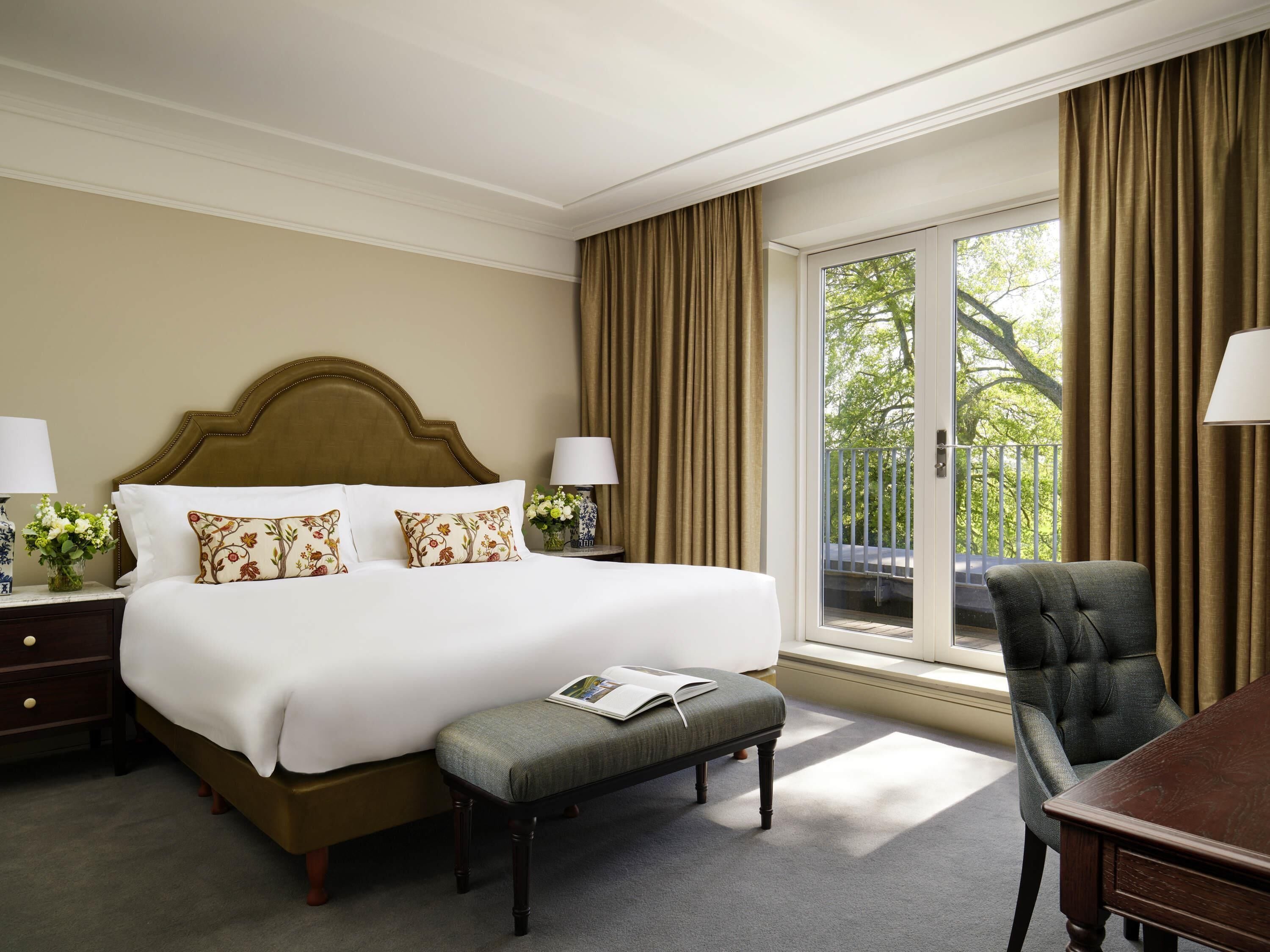 A hotel room with a large bed, a desk, a chair, a window with curtains, and a view of greenery outside.