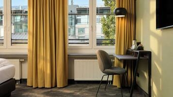Ein Hotelzimmer mit einem Bett, einem Schreibtisch mit Stuhl, einer Lampe und Blick auf Gebäude durch große Fenster.