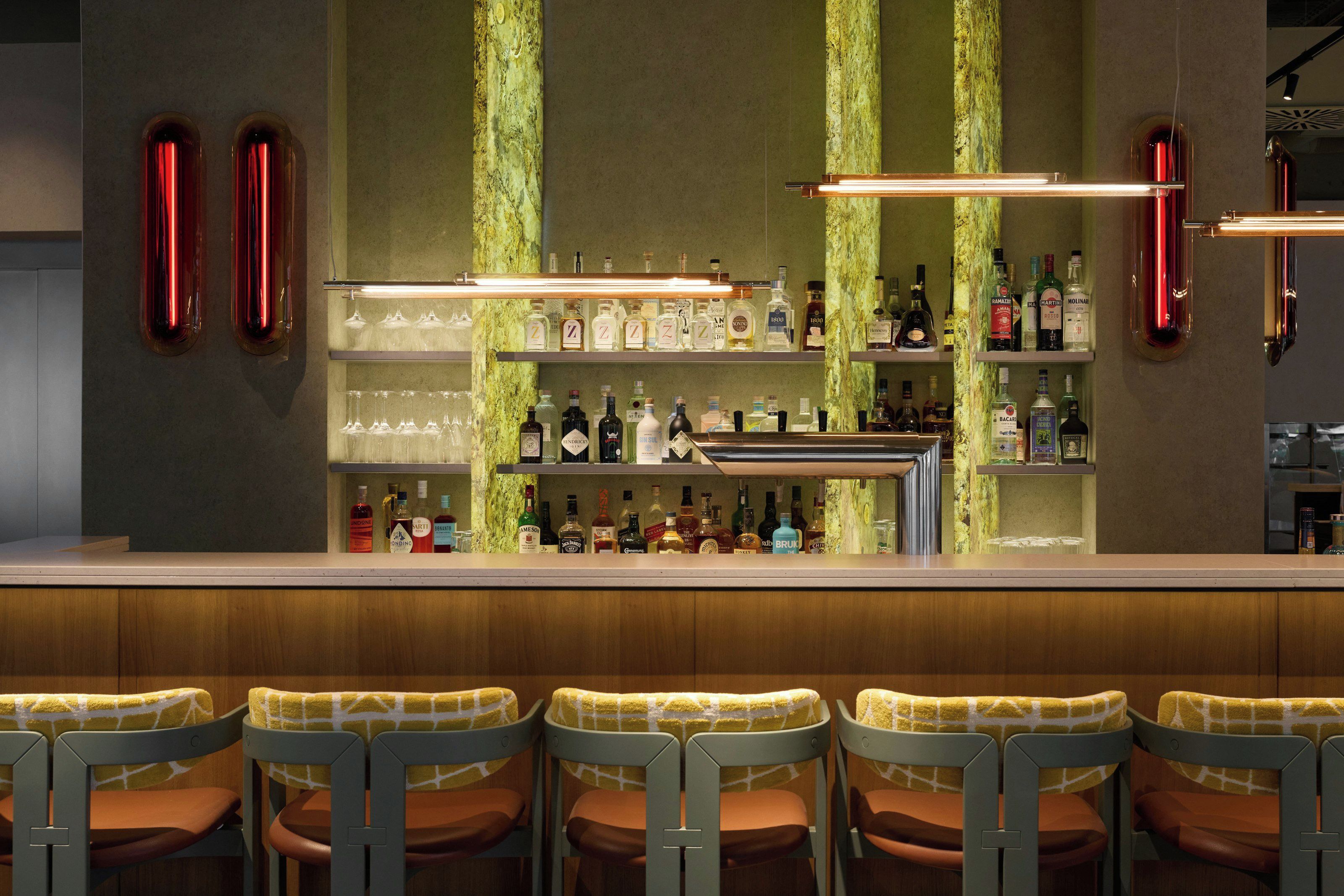 Uma área de bar com um balcão comprido, várias prateleiras cheias de garrafas de bebidas e uma fileira de banquetas com almofadas estampadas.