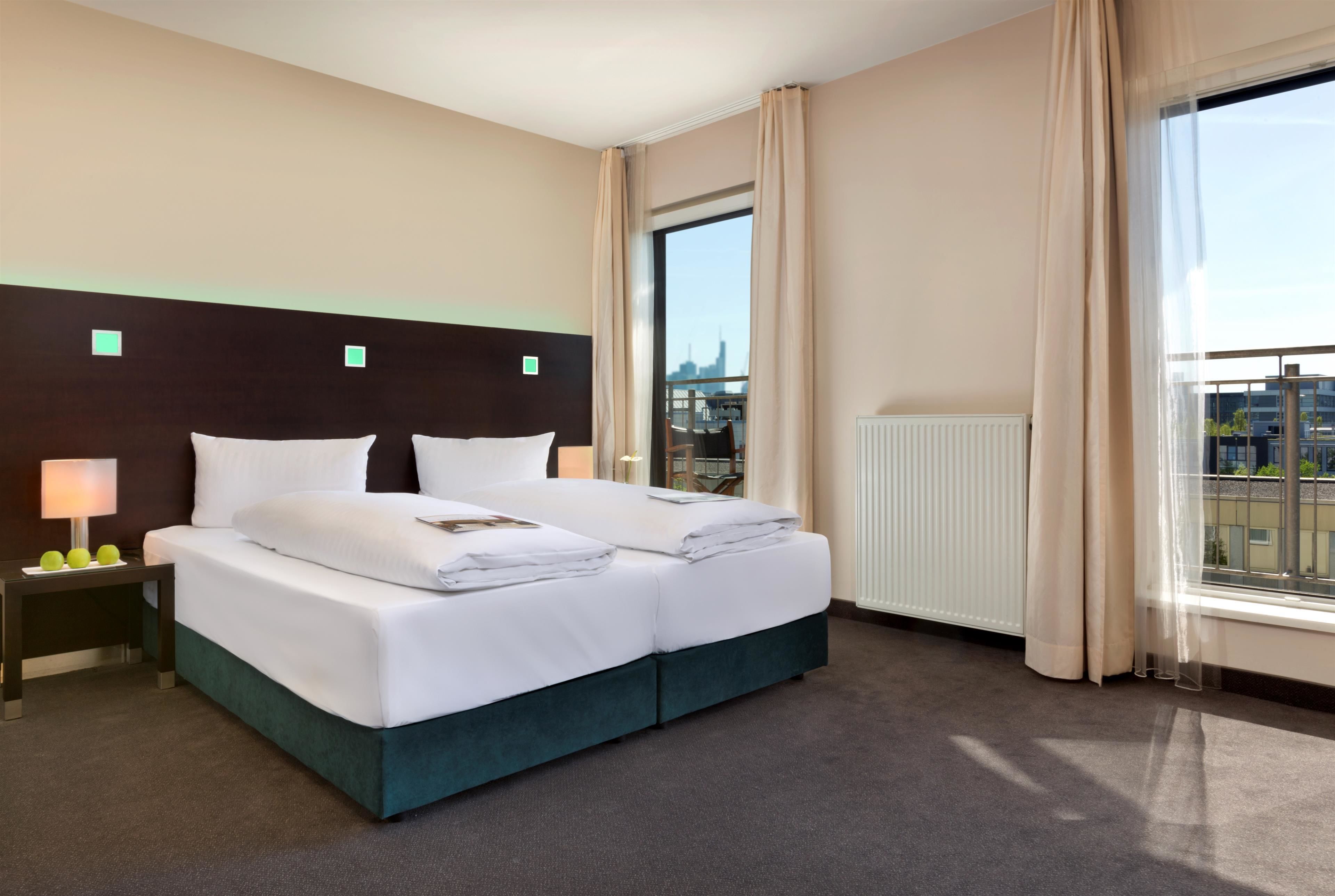 A hotel room with a large bed, a nightstand with a lamp, and a view of the city through a window.