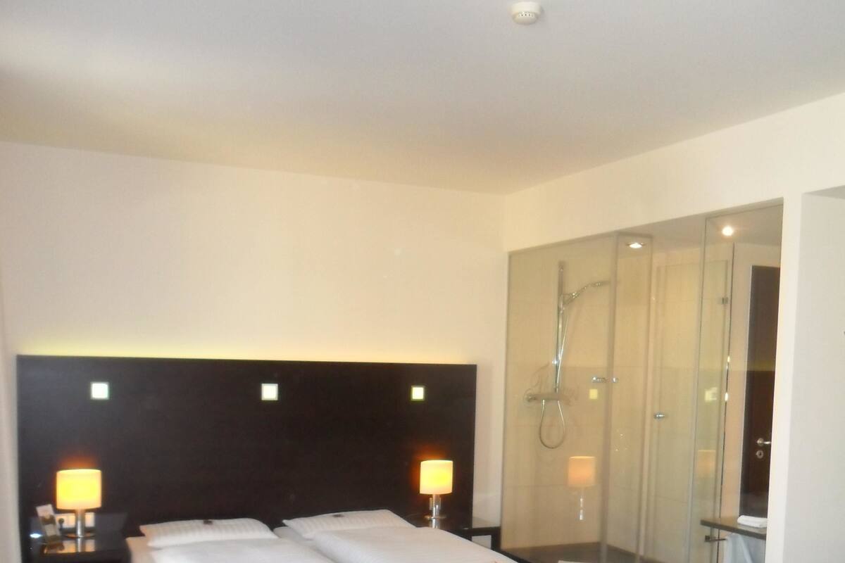 Een hotelkamer met een bed, twee bedlampen, een douche met glazen wanden, een houten tafel met appels en een bruine leren bank.