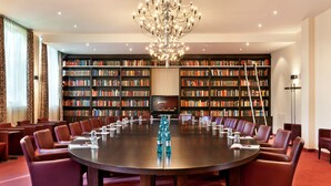 Un salón de conferencias con una mesa redonda rodeada de sillas, una araña y estanterías al fondo.
