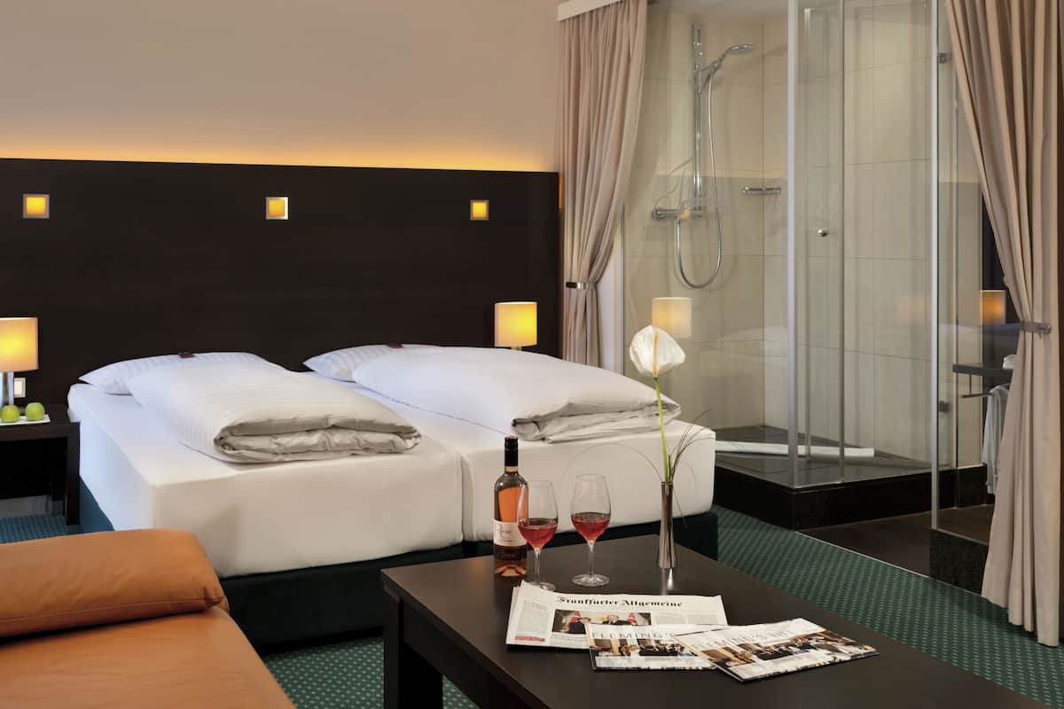 Hotelkamer met een bed, een douche, een tafel met wijnglazen en een fles, en een tijdschrift.