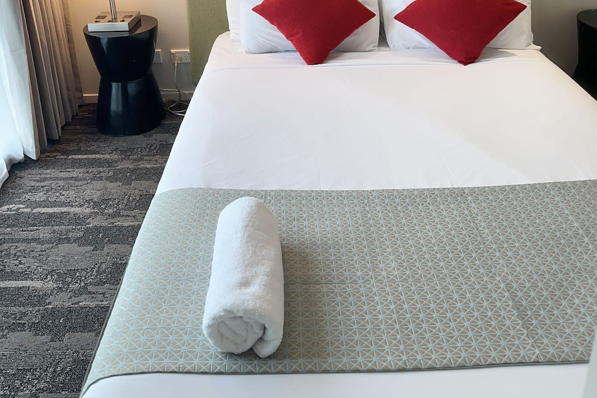 A hotel room with a neatly made bed, a headboard, a framed artwork above the bed, a bedside lamp, and a patterned rug.