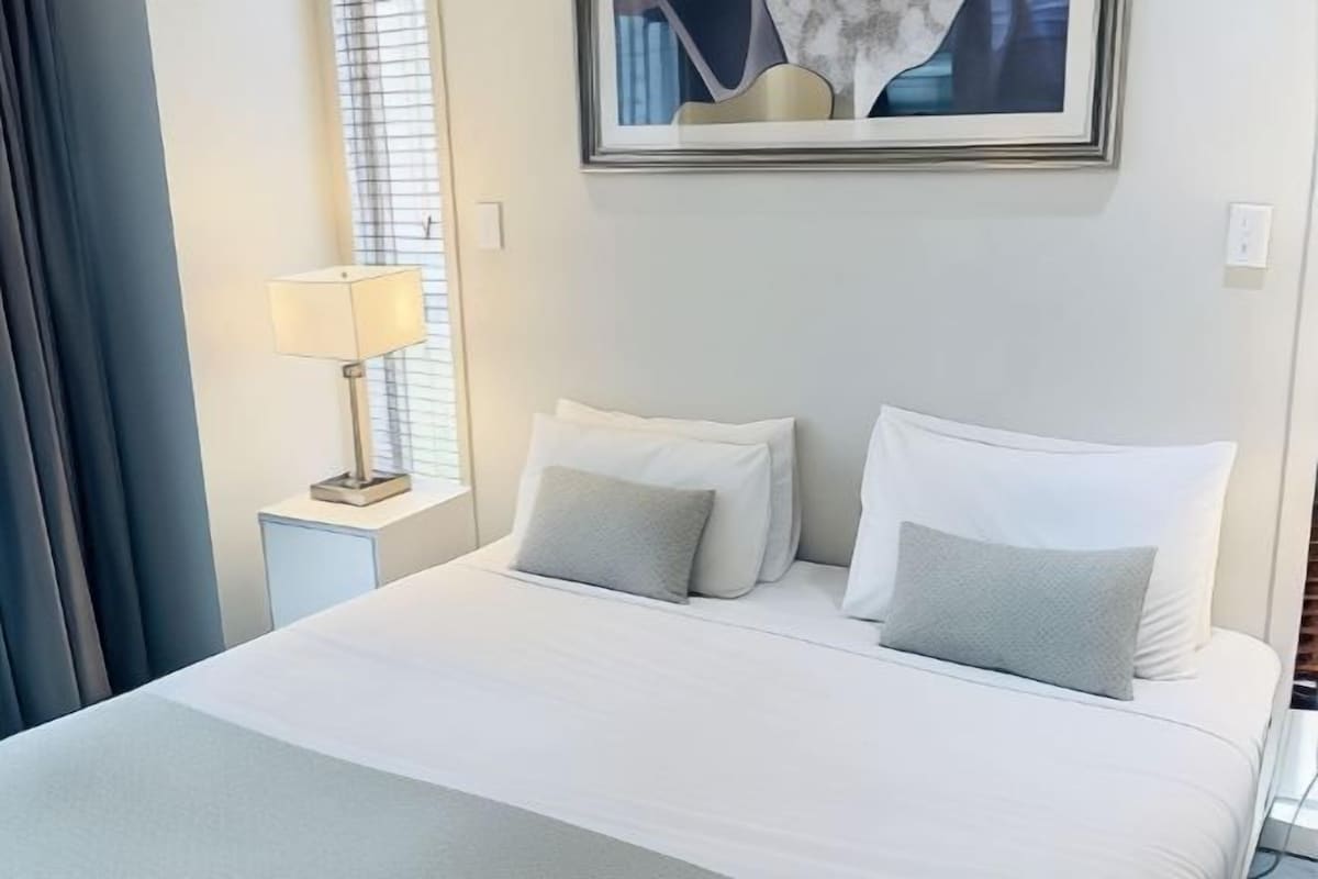 A hotel room with a bed, pillows, a nightstand with a lamp, and a framed picture on the wall.