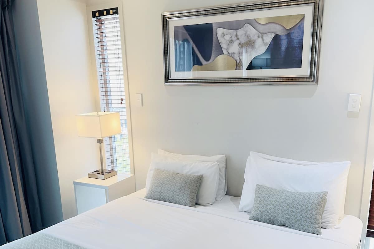 A neatly made bed with white linens and a grey blanket, a bedside lamp, and a framed abstract painting on the wall.