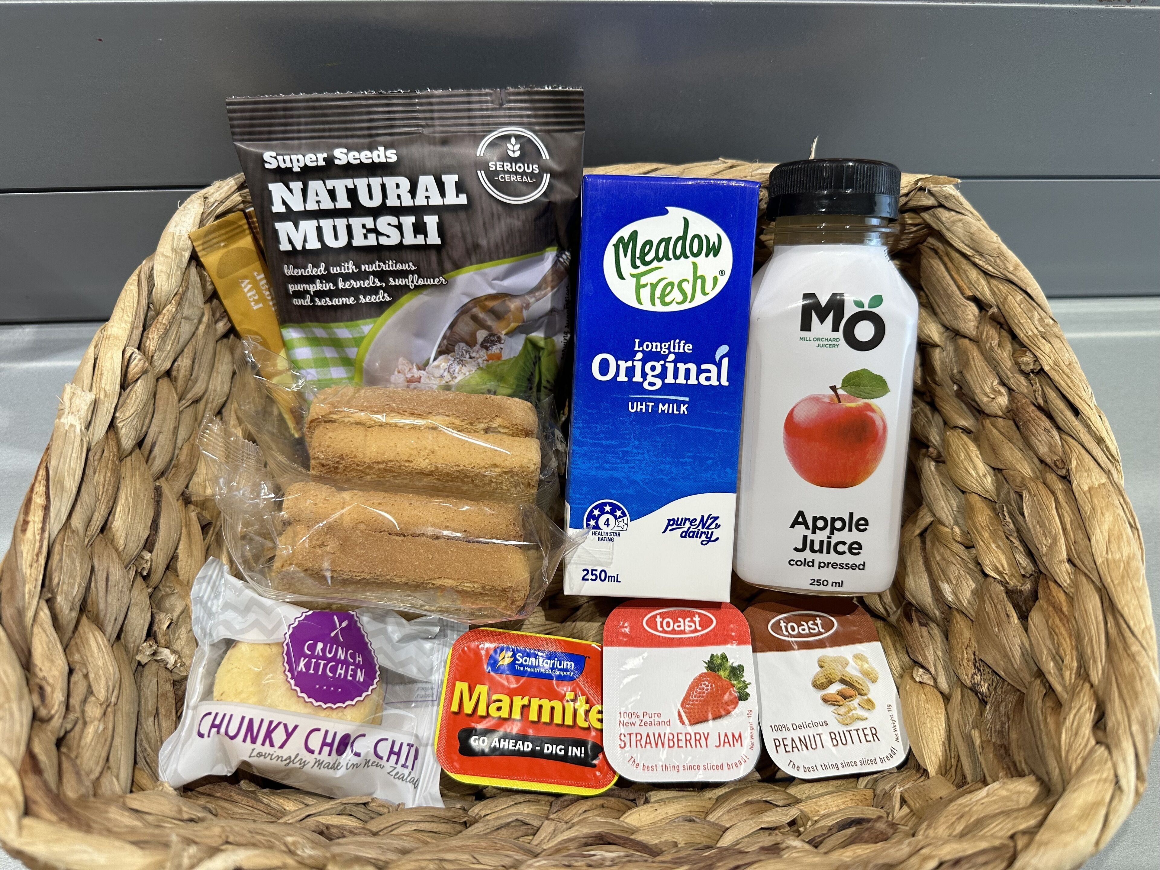 A basket containing various food items including a package of natural muesli, Meadow Fresh milk, apple juice, and snacks like chunky chocolate chip cookies and marmite.
