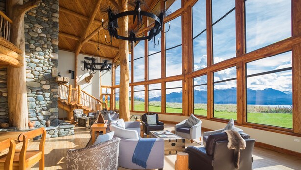 A spacious living area with a stone wall, wooden ceiling, and large windows offering a mountain view. - Fiordland Lodge (Te Anau)