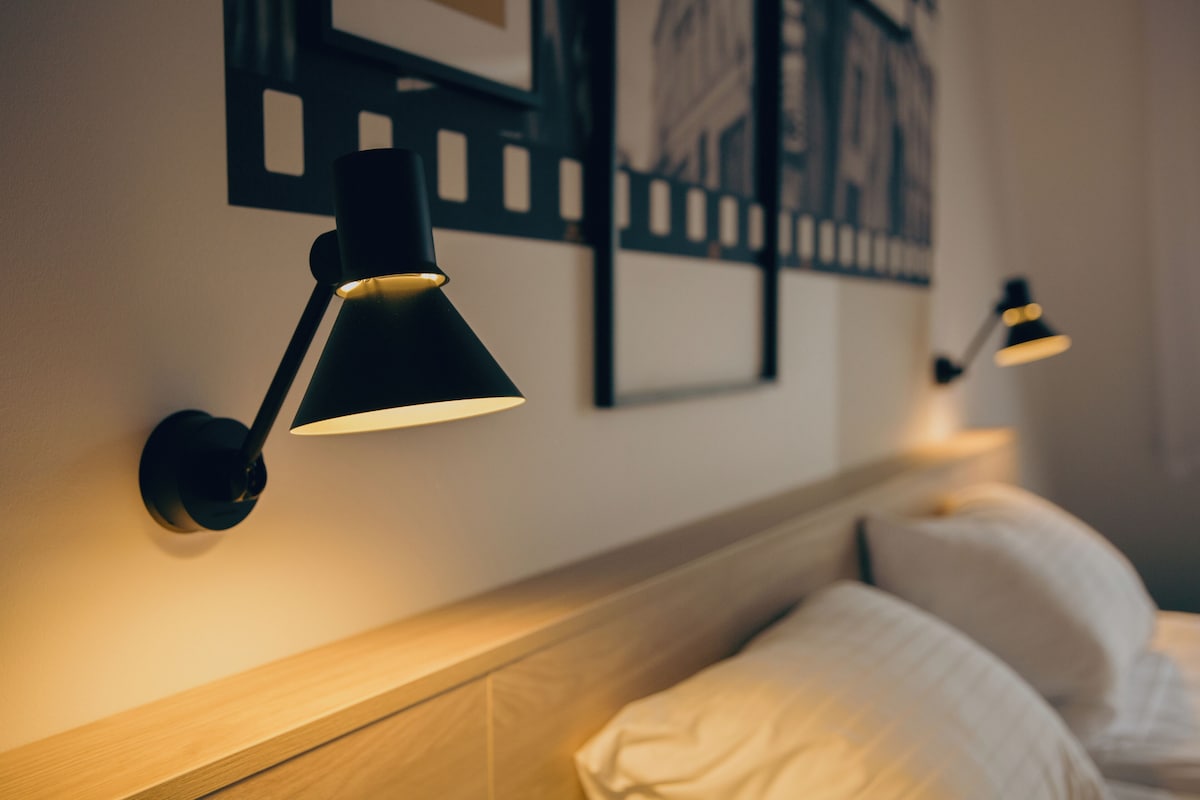 Una habitación de hotel con cabecera de madera, una cama con ropa blanca y dos lámparas fijadas a la pared.