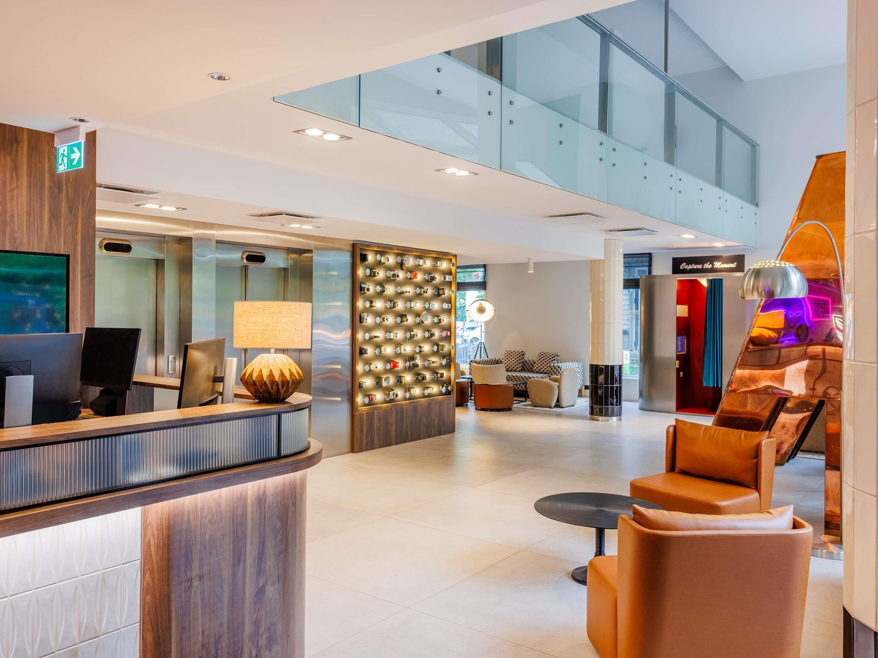 A modern hotel lobby with a reception desk, seating area, and a glass staircase.
