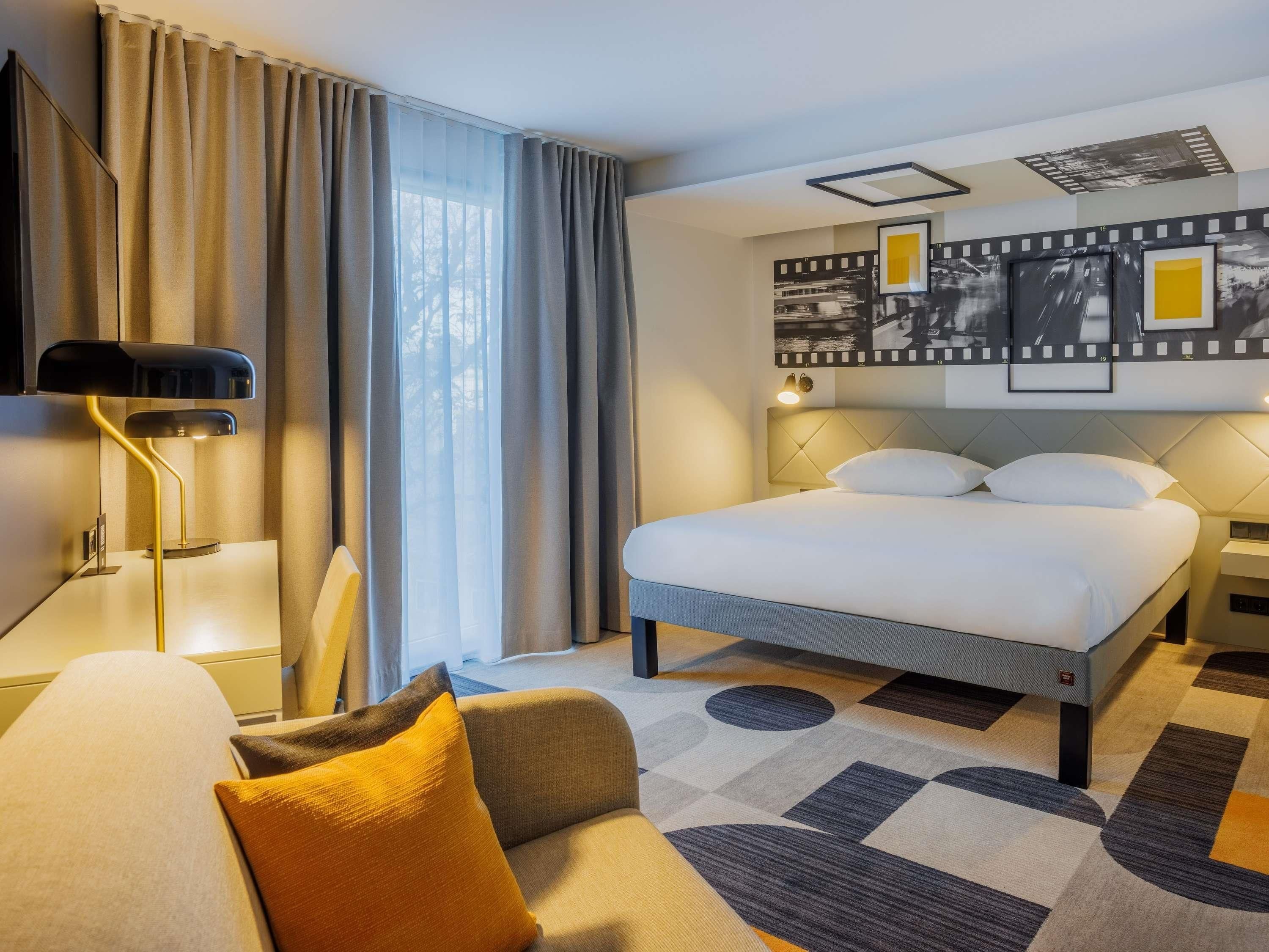 A hotel room with a bed, a desk, a chair, a lamp, and a wall with framed pictures.