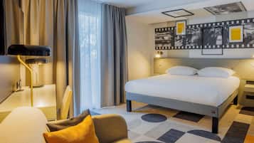 A hotel room with a bed, a desk, a chair, a lamp, and a wall with framed pictures.