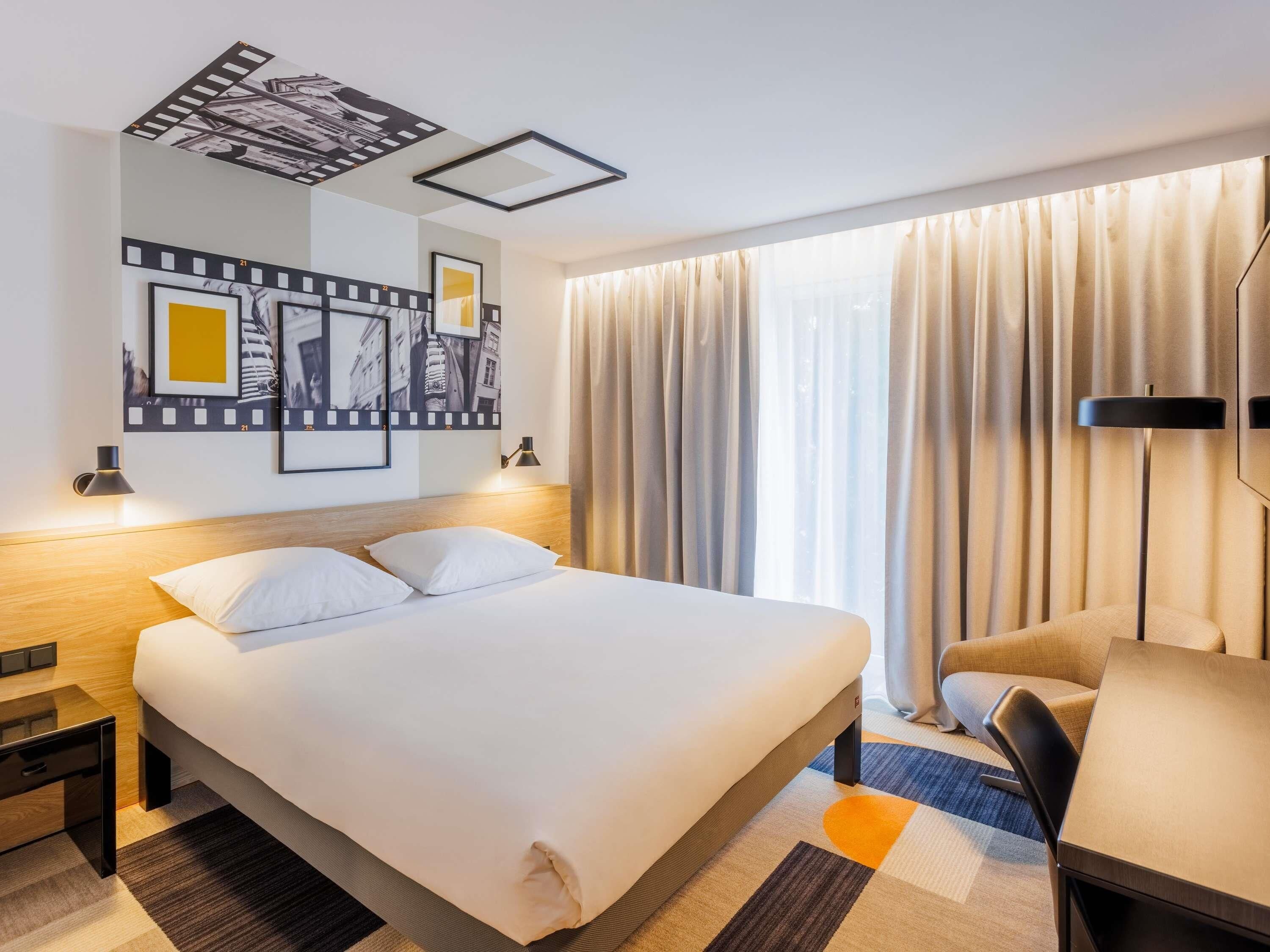 Habitación de hotel con cama, escritorio, silla, un ventanal con cortinas y decoración en la pared.