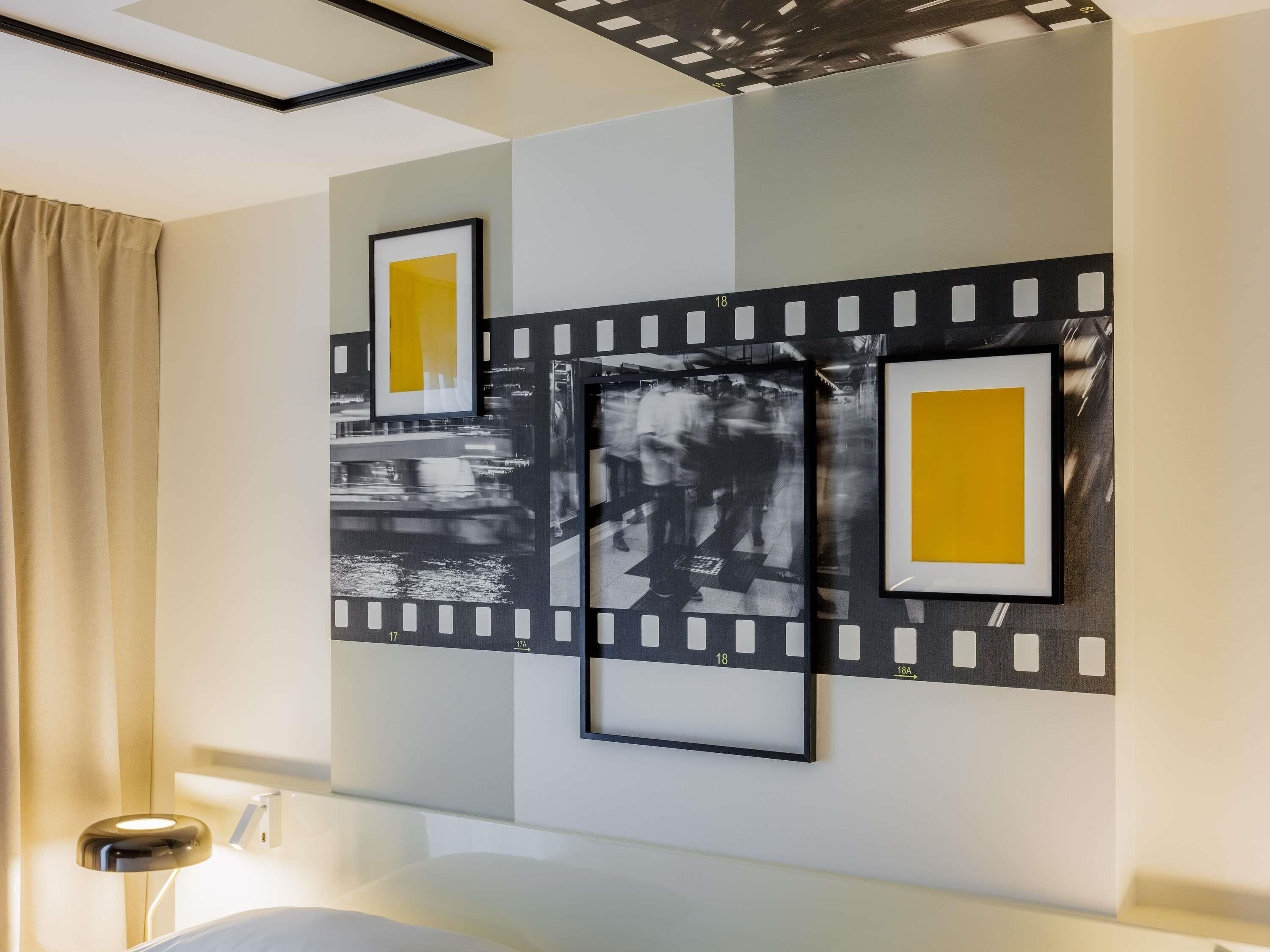 A hotel room with a bed, a bedside table with a lamp, and a wall with framed pictures.