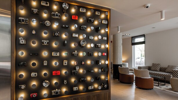 A wall with numerous cameras displayed in a grid pattern, a modern sofa, and a wooden floor in a well-lit room. - ibis Styles Tallinn (Tallinn)