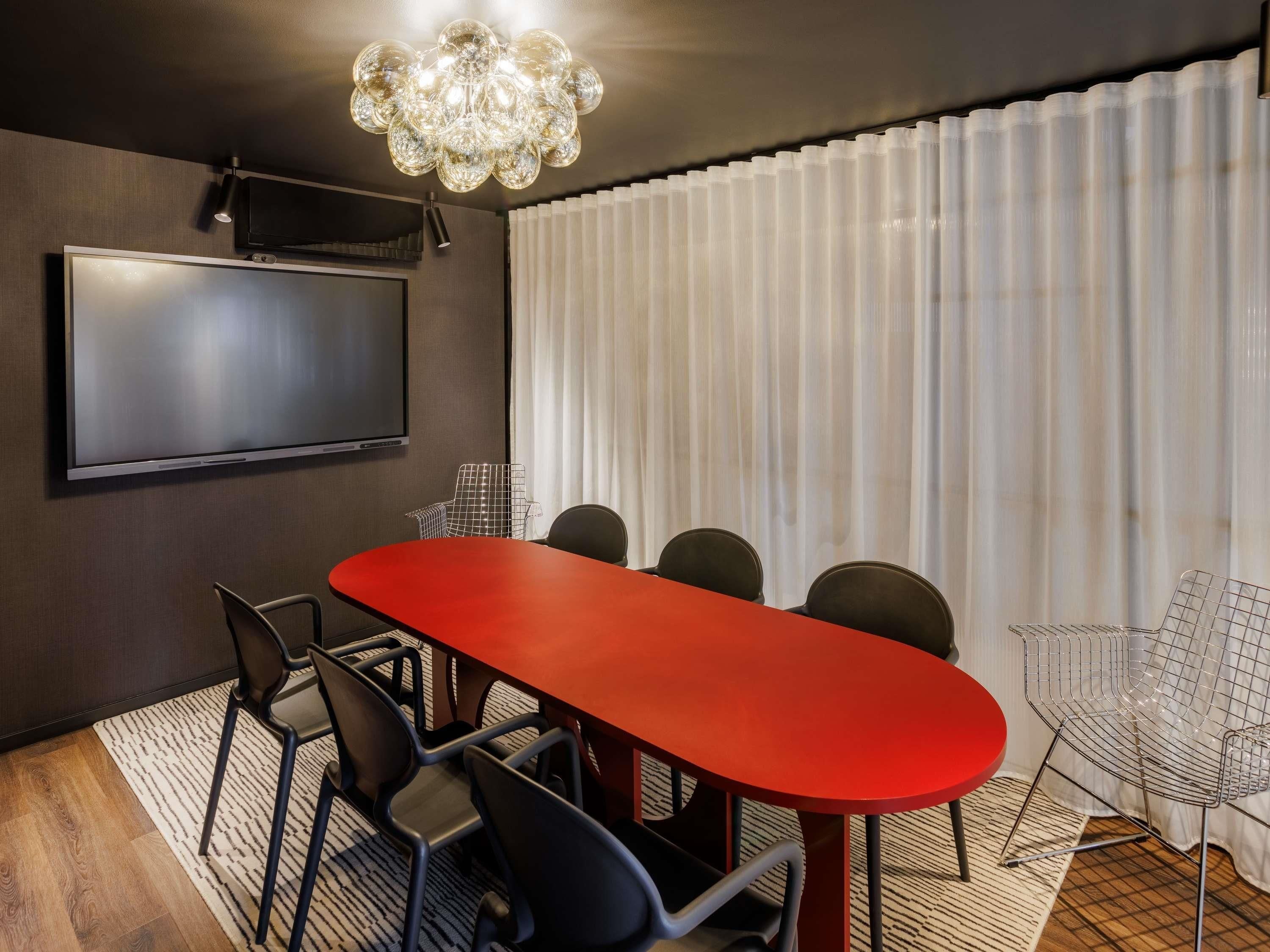 Une salle de conférence avec une table rouge, des chaises noires, un téléviseur et un lustre moderne.