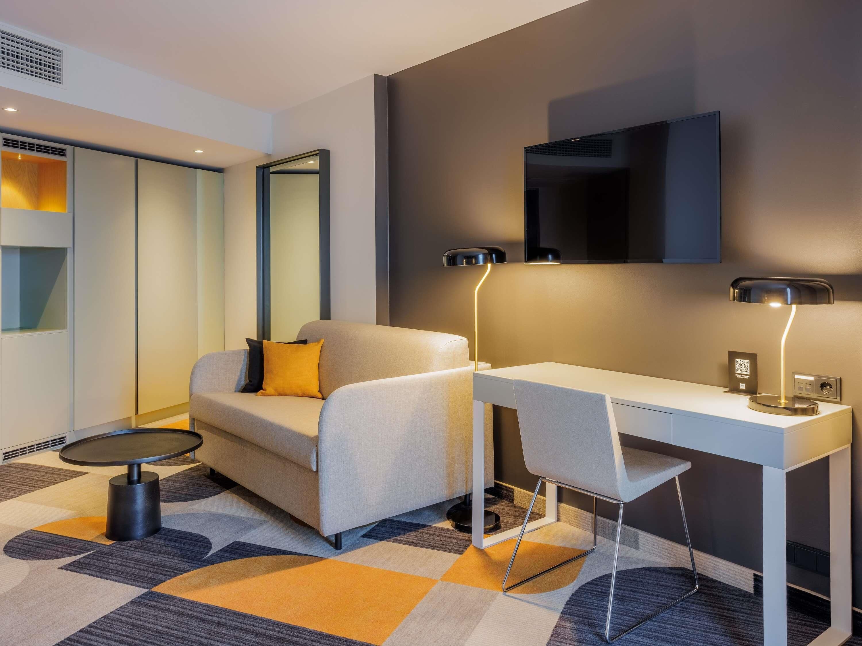 A modern hotel room with a sofa, a desk, a round table, and a flat-screen TV mounted on the wall.