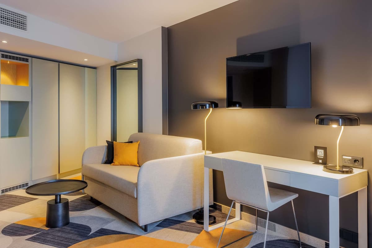 A modern hotel room with a sofa, a desk, a round table, and a flat-screen TV mounted on the wall.