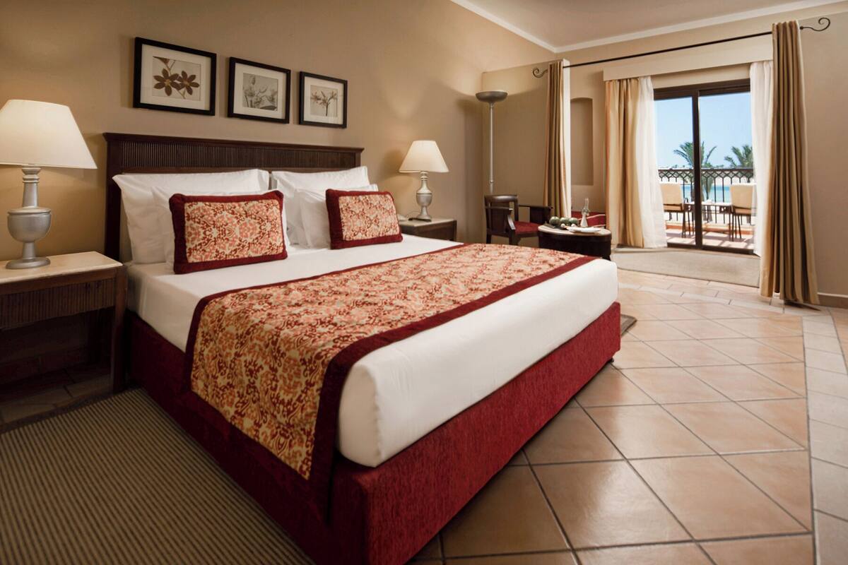 A hotel room with a large bed, two bedside tables with lamps, a view of the balcony, and framed pictures on the wall.