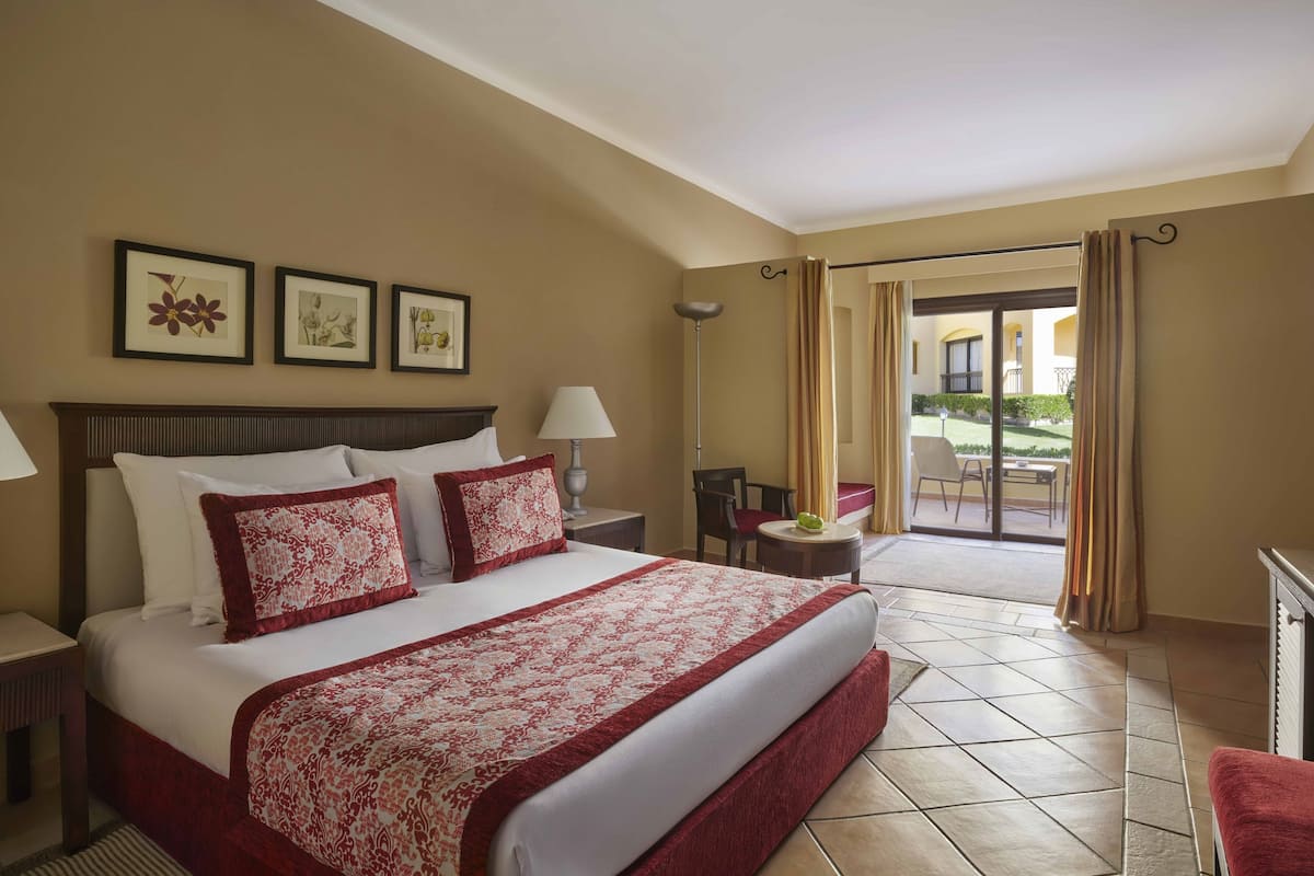 A hotel room with a large bed, a desk, a chair, and a view of the outdoor area through a sliding door.
