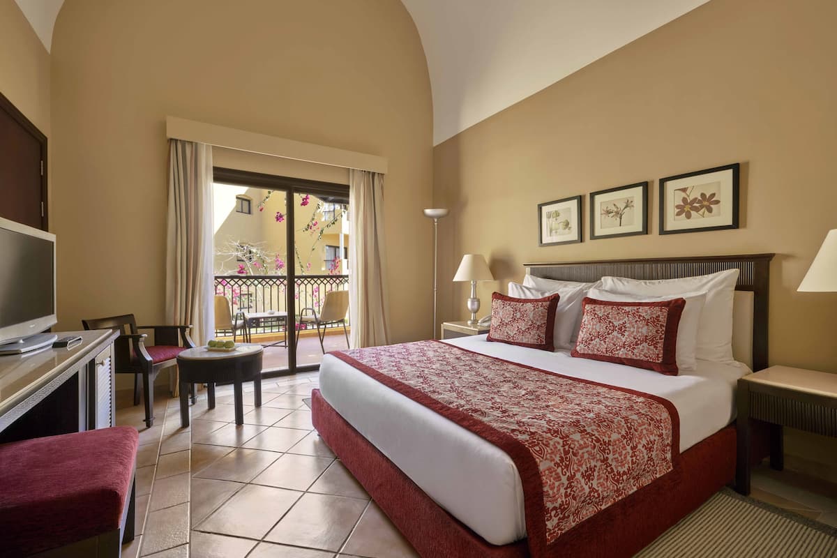 A hotel room with a large bed, a desk with a TV, a chair, and a balcony with a view of buildings.
