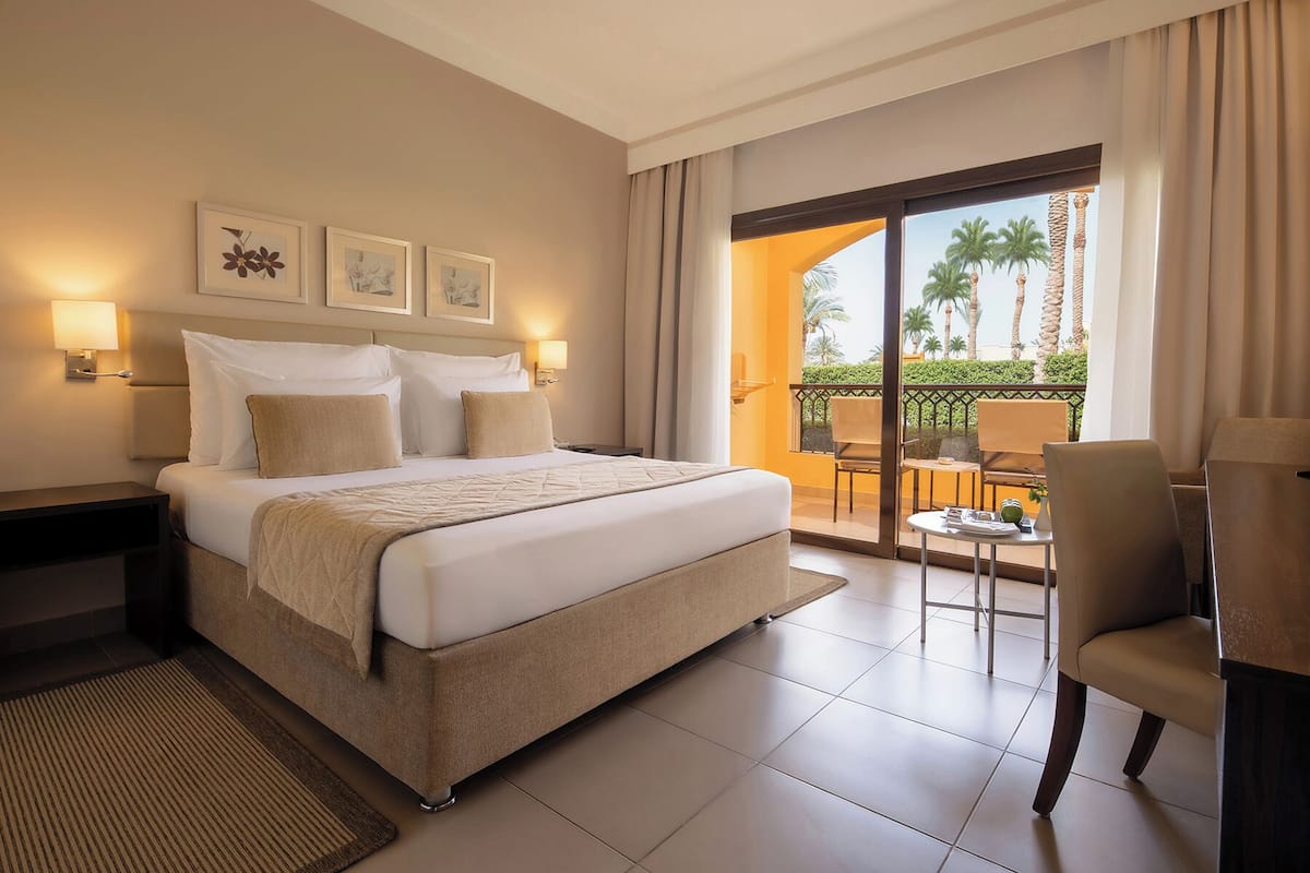 A hotel room with a large bed, a balcony with chairs, and a view of palm trees.