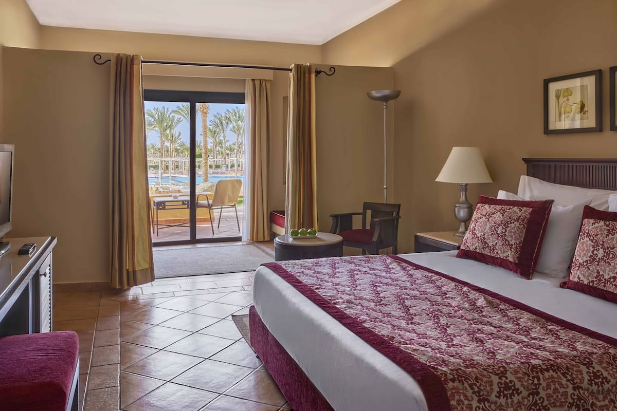 A hotel room with a bed, a desk, a chair, a lamp, and a view of a pool and palm trees.
