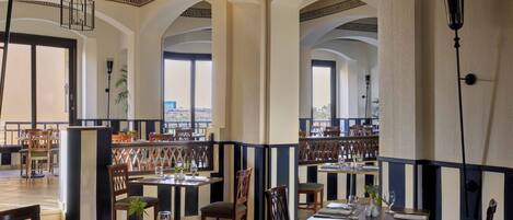 A spacious restaurant with high ceilings, arched windows, and a view of the cityscape. Tables are neatly arranged with chairs, and the decor includes patterned columns and a striped wall.
