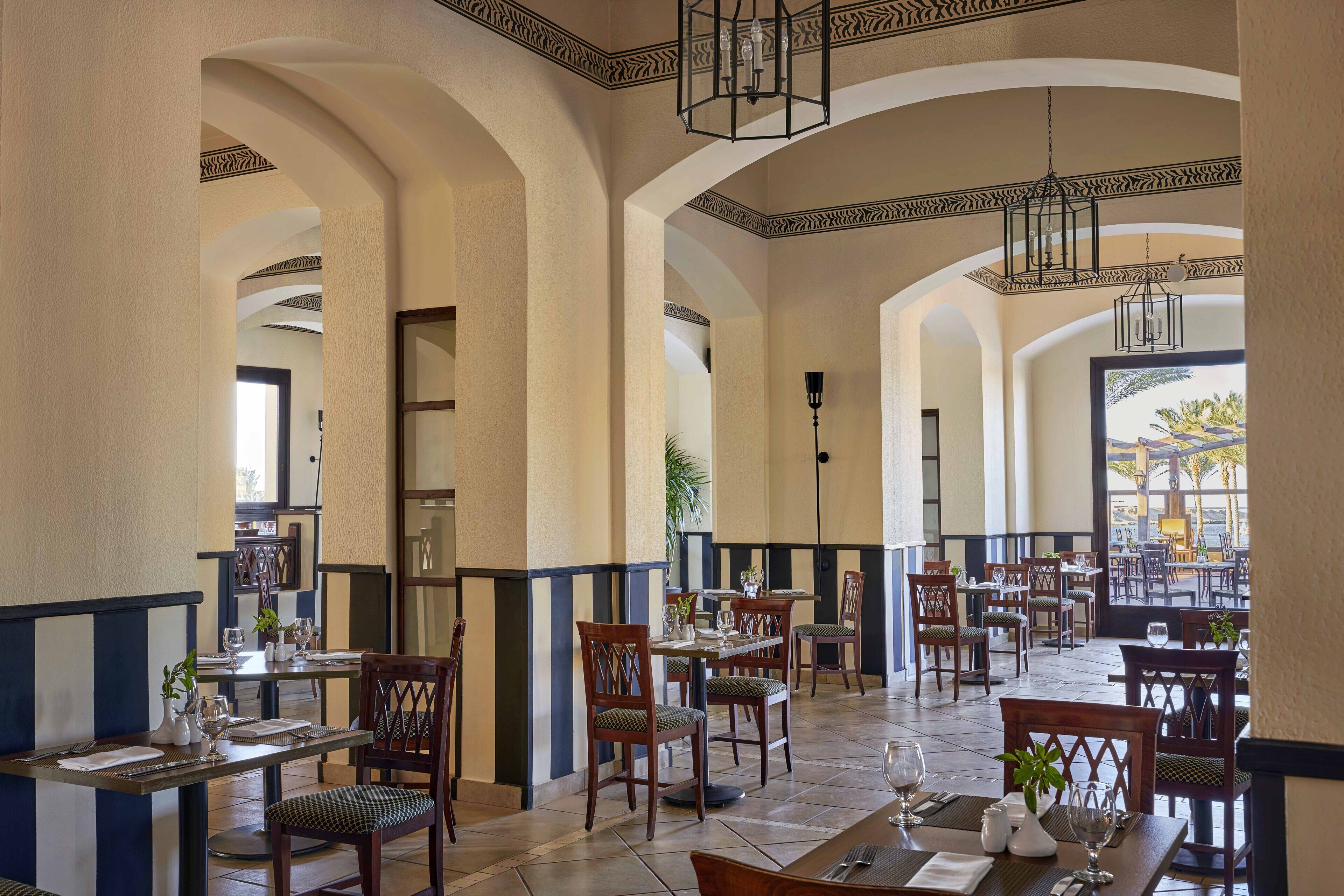 A spacious dining area with high ceilings and arched doorways, featuring multiple tables and chairs, and a view of the outdoors through large windows.
