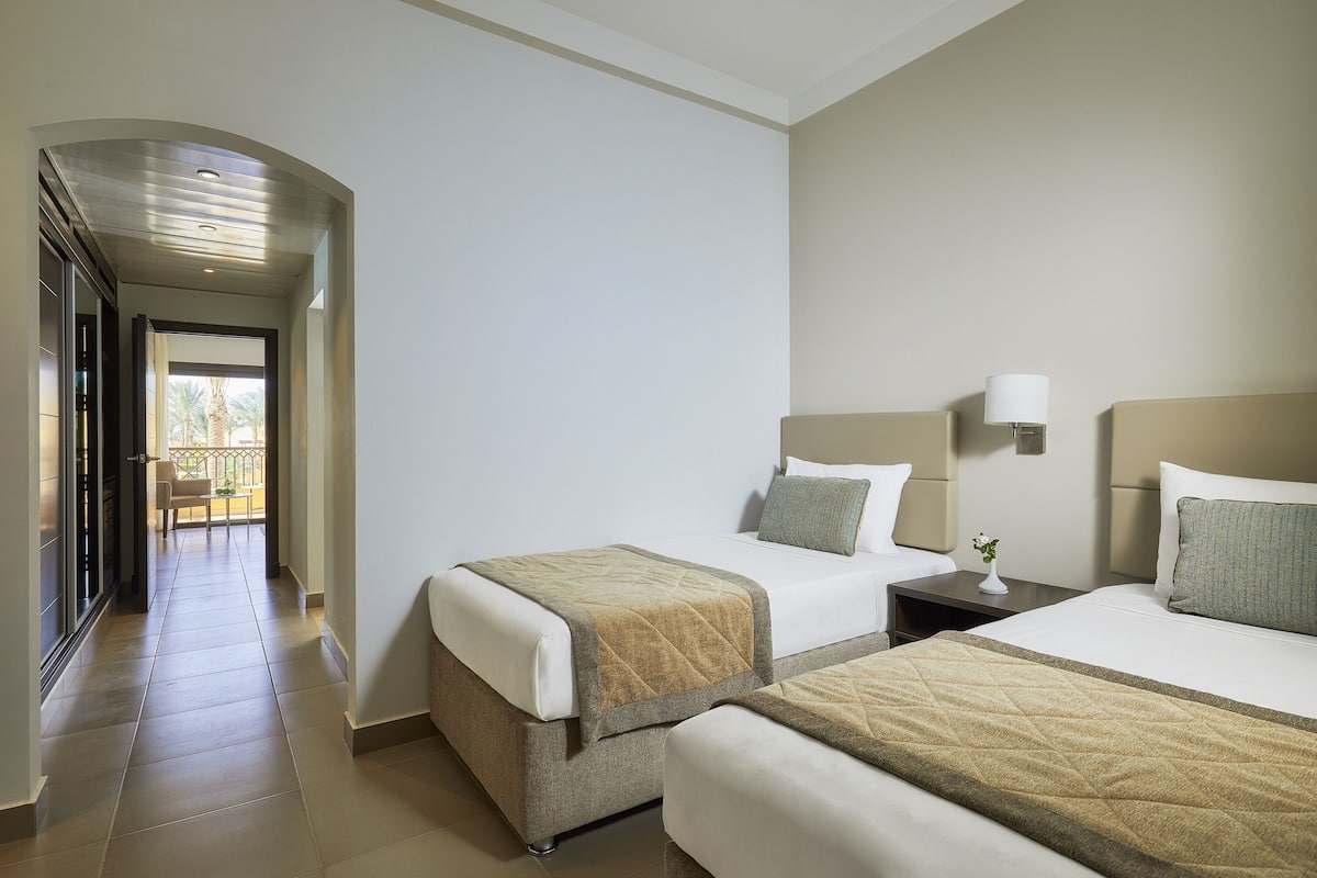 A hotel room with two beds, a nightstand, a lamp, and a view of a balcony.